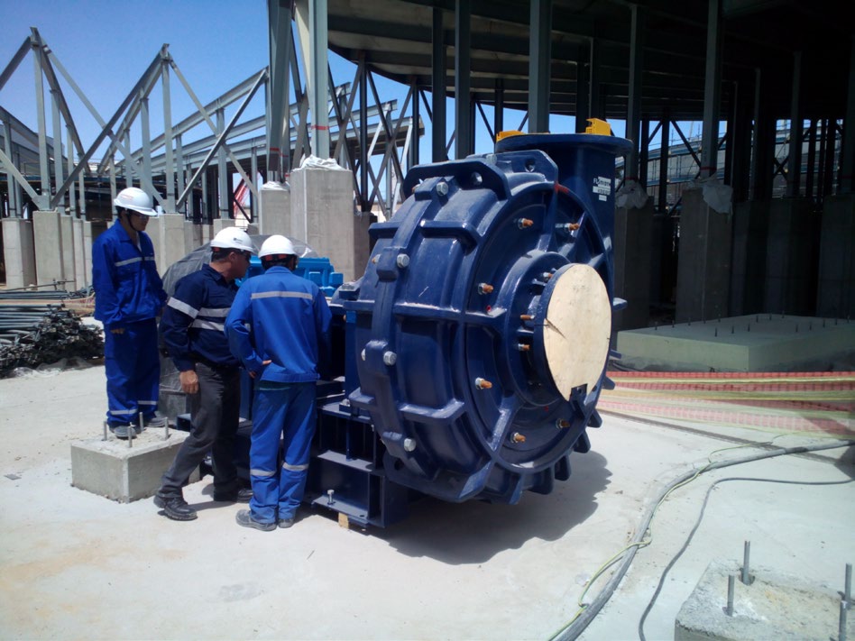 Three construction workers in hard hats and blue overalls examining a large, dark blue industrial machine or pump at a construction site with structural steel in the background.